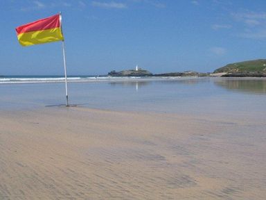 A sandy beach with a red and yellow flag on a pole and a lighthouse on a rocky island in the distance at Godrevy Cottage in Hayle