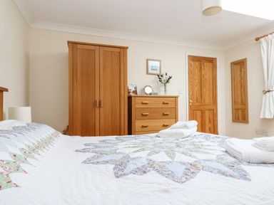 A bedroom with a bed covered by a patterned quilt folded towels on the bed wooden wardrobe and dresser a door and a wall mirror at Godrevy Barn in Hayle