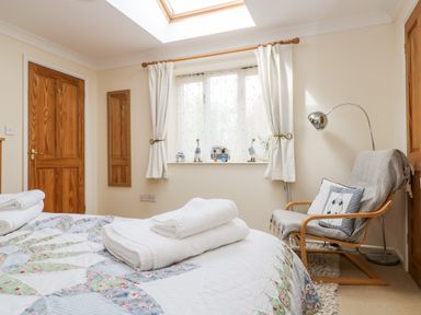 A bedroom with a bed covered in a patterned quilt and stacked towels a wooden chair with a cushion a floor lamp and a window with curtains at Godrevy Barn in Hayle