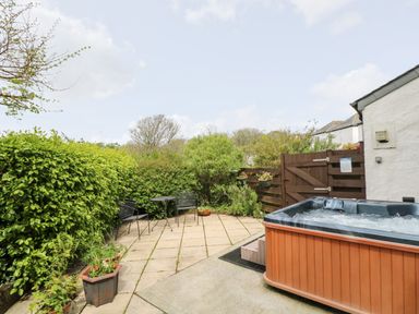 An outdoor patio area with a hot tub black metal chairs and table surrounded by green bushes at Godrevy Barn in Hayle