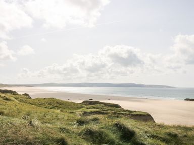 A sandy beach with grassy dunes in the foreground and calm sea under a cloudy sky at Godrevy Barn in Hayle