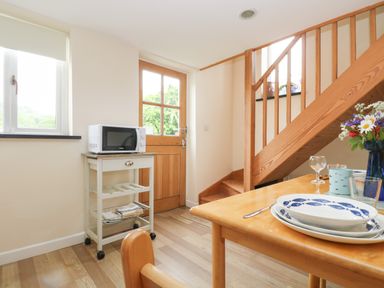 A kitchen area with a microwave on a cart near a wooden door and a wooden staircase next to a table with dishes and flowers at Little Hamlin in Rilla Mill Nr Callington