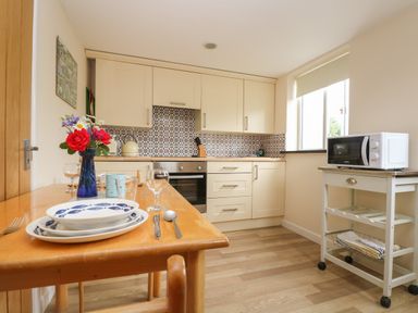 A kitchen with a wooden dining table set with dishes and flowers and a microwave on a white cart by the window at Little Hamlin Rilla Mill Nr Callington