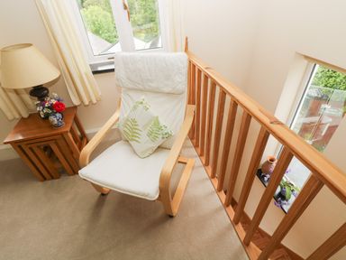 A white cushioned chair with a leaf-patterned pillow next to a wooden side table with a lamp and flowers near windows and wooden railing at Little Hamlin in Rilla Mill Nr Callington