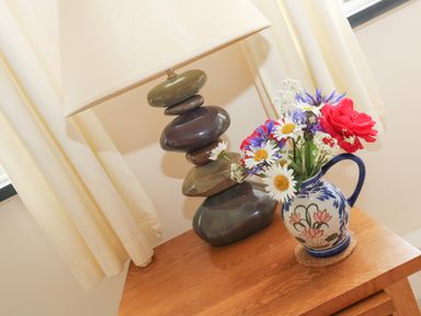 A table lamp and a jug with flowers on a wooden surface by a window with curtains at Little Hamlin in Rilla Mill Nr Callington