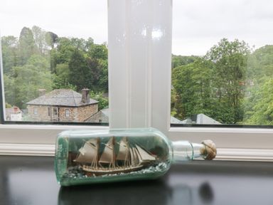 A ship in a glass bottle on a windowsill with trees and a house visible outside at Little Hamlin in Rilla Mill Nr Callington