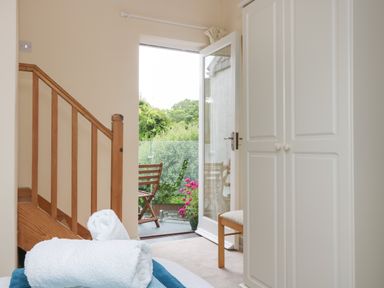 A room with a wooden staircase, white wardrobe, open door leading to a balcony with a wooden chair and pink flowers at Little Hamlin in Rilla Mill Nr Callington