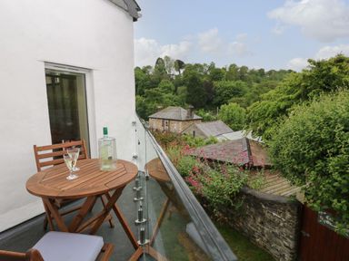 A balcony with a wooden table and chairs and a bottle with two glasses overlooking greenery and rooftops at Little Hamlin in Rilla Mill Nr Callington