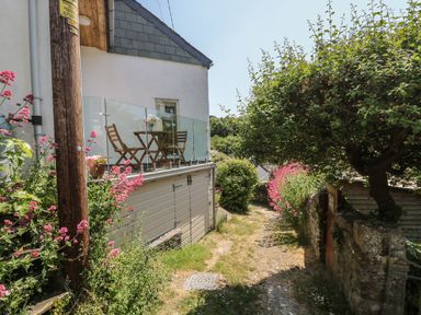 A garden path beside a house with a balcony with a table and chairs and flowering plants at Little Hamlin in Rilla Mill Nr Callington