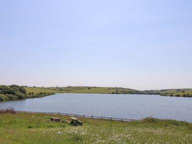 A lake with picnic tables on grassy shore and hills in the background at Little Hamlin in Rilla Mill Nr Callington