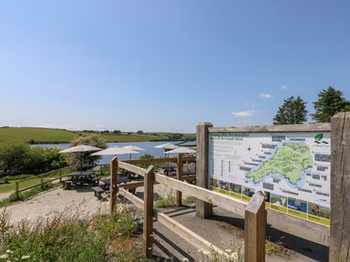 An outdoor seating area with umbrellas and picnic tables near a lake with a map board on a wooden fence at Little Hamlin in Rilla Mill Nr Callington