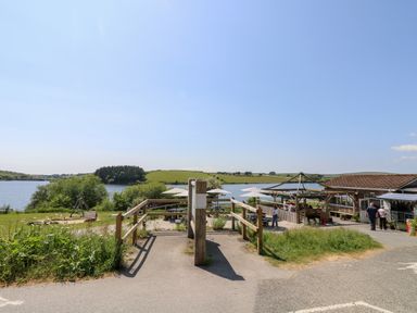 An outdoor seating area with tables and umbrellas near a lake and grassy fields at Little Hamlin in Rilla Mill Nr Callington