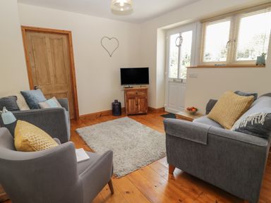 A living room with grey sofas and armchair around a rug a wooden door and a small TV on a wooden stand at Honeysuckle Cottage in Helston