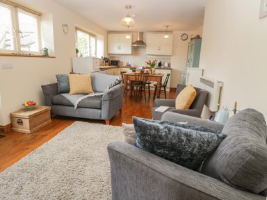 A living room with grey sofas, a wooden dining table and chairs, kitchen cabinets, and wooden floor at Honeysuckle Cottage in Helston