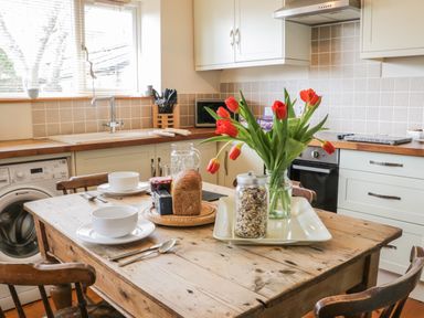 A kitchen with a wooden table set with bowls bread jar of cereal and red flowers in Helston