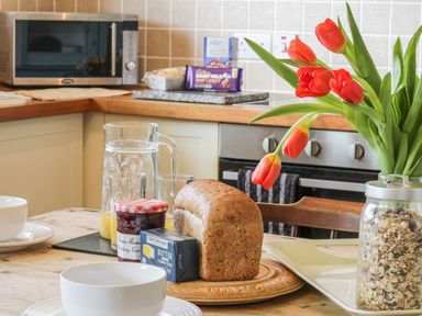 A kitchen table with bread jam butter a jar of cereal a pitcher with glasses and red tulips at Honeysuckle Cottage in Helston