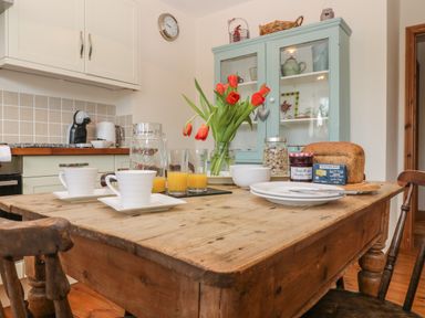 A kitchen table with cups plates orange juice bread butter and flowers at Honeysuckle Cottage in Helston