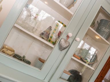 A glass cabinet with glass bottles ceramic containers a metal bucket and decorative items at Honeysuckle Cottage in Helston