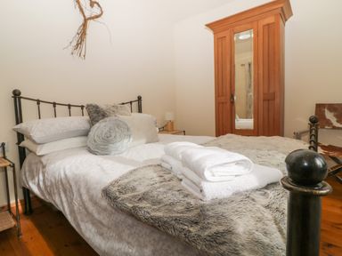 A bedroom with a bed with white linens and gray pillows a wooden wardrobe with a mirror and a chair at Honeysuckle Cottage in Helston