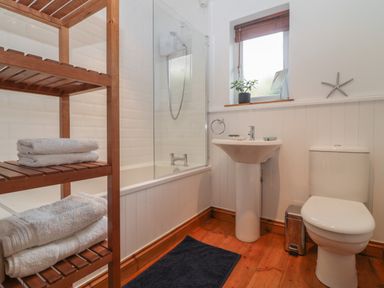 A bathroom with a bathtub and shower a sink a toilet a wooden towel rack with white towels and a small window at Honeysuckle Cottage in Helston