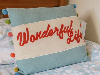 A decorative pillow with colorful pom poms and the words Wonderful life on a bed with floral sheets at Honeysuckle Cottage in Helston
