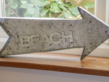 A metal arrow sign with the word beach on a wooden windowsill with greenery outside at Honeysuckle Cottage in Helston