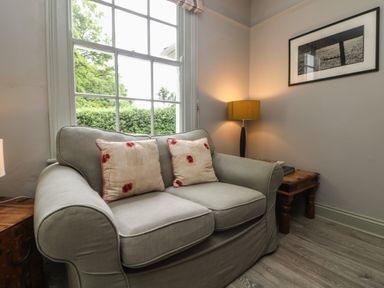 A seating area with a two-seater sofa with floral cushions beside a window and wooden tables with a lamp and a framed picture at Penwarne Lodge in Mawnan Smith
