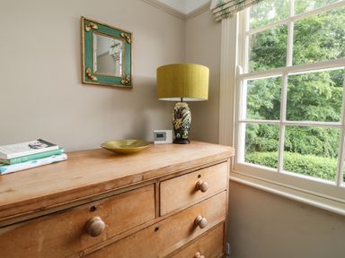 A wooden chest of drawers with books a decorative plate and a lamp next to a window at Penwarne Lodge in Mawnan Smith