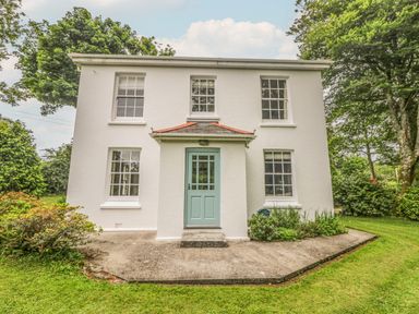 A white two-story house with a blue front door and four windows surrounded by trees and grass at Penwarne Lodge in Mawnan Smith