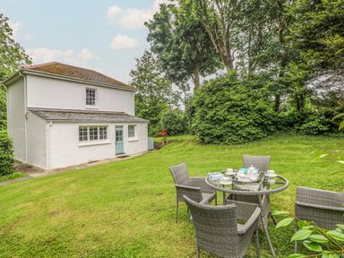 A small white house with a blue door and four grey wicker chairs around a glass table on a lawn at Penwarne Lodge in Mawnan Smith