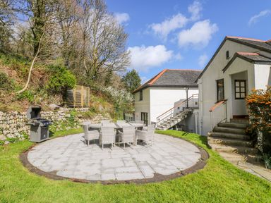An outdoor patio with a round table and chairs a barbecue grill and stairs leading to a white building at Carne Mill in Manaccan