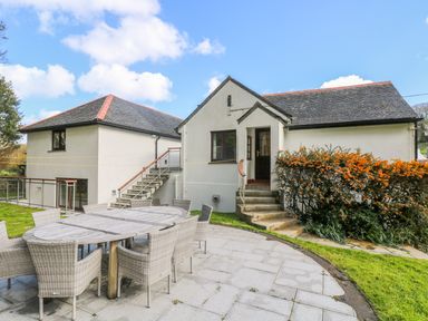 An outdoor patio with a large wooden table and wicker chairs outside a white building with stairs and orange flowering bushes at Carne Mill in Manaccan