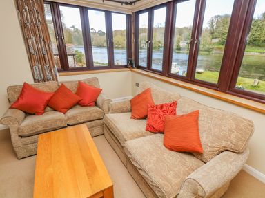 A living room with two beige patterned sofas with red cushions a wooden coffee table and large windows overlooking a river at Carne Mill in Manaccan
