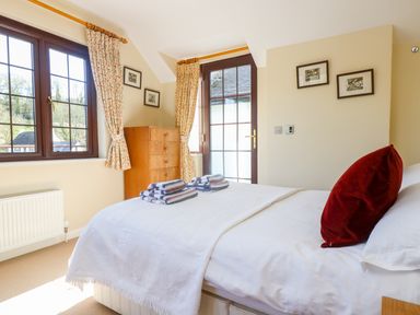 A bedroom with a bed, folded towels, a chest of drawers, two windows with curtains, and framed pictures on the walls at Carne Mill in Manaccan