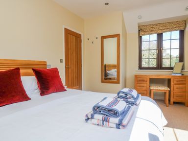 A bedroom with a bed with red pillows folded towels on the bed a wooden desk with a stool and a window at Carne Mill in Manaccan