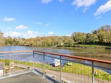 A balcony overlooking a river with a boat on the shore and trees in the background at Carne Mill in Manaccan