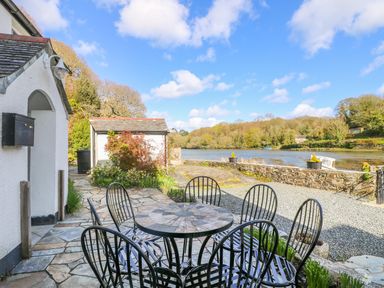 An outdoor patio with a round stone table and black metal chairs overlooking a river and trees at Carne Mill in Manaccan