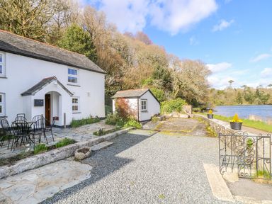 A white house with a small patio and metal table and chairs near a gravel driveway by a lake at Carne Mill in Manaccan