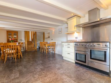 A kitchen with a stainless steel oven and hood wooden dining tables and chairs with stairs in the background at Carne Mill in Manaccan