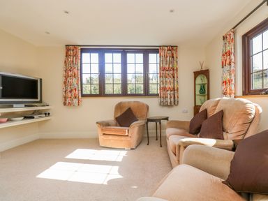 A sitting room with beige armchairs a television on a wall shelf and windows with floral curtains at Carne Mill in Manaccan