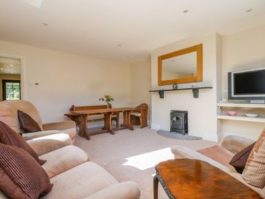 A living room with beige armchairs a wooden table and bench a stove under a wall mirror and a television on shelves at Carne Mill in Manaccan
