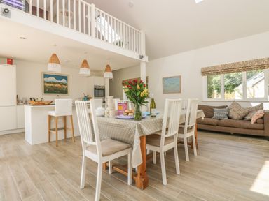 A dining area with a table and chairs next to a kitchen with stools and hanging lights in a room with a sofa by the window at Little Chycoose in Truro