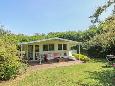 A wooden garden shed with a covered porch, outdoor seating, a green kayak, and a barbecue grill in a garden at Little Chycoose in Truro