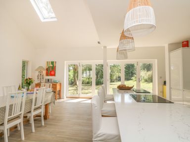 A kitchen and dining area with a long white island and chairs near glass doors showing a garden at Little Chycoose in Truro