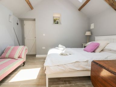 A bedroom with a bed with white bedding and a pink pillow a pink and white patterned chaise lounge a wooden floor and wooden beams on the ceiling at Little Chycoose in Truro
