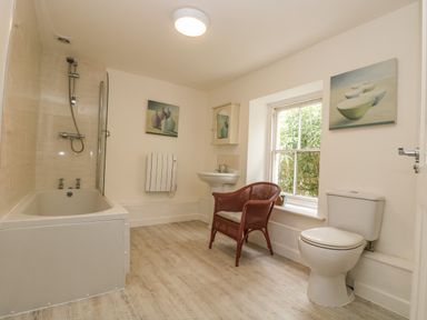 A bathroom with a bathtub shower sink toilet and wicker chair by a window at Little Chycoose in Truro