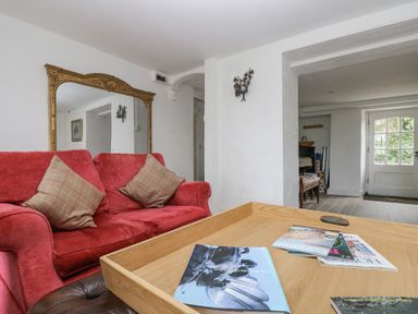 A room with a red sofa with two plaid cushions a large ornate mirror a wooden tray with magazines and a view into a hallway with a white door at Little Chycoose in Truro