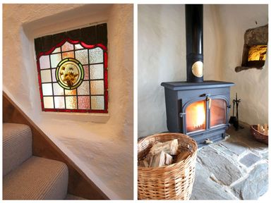 A stained glass window next to carpeted stairs and a wood burning stove with a basket of firewood on a stone floor at Rose Cottage in Manaccan
