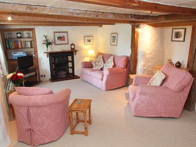 A living room with exposed wooden beams pink upholstered sofa and chairs a fireplace and wooden furniture at Rose Cottage in Manaccan