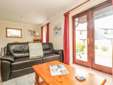 A living room with a black leather sofa a wooden coffee table with an open magazine and a tin and red curtains over wooden framed glass doors at Cuckoo's Cottage in Falmouth